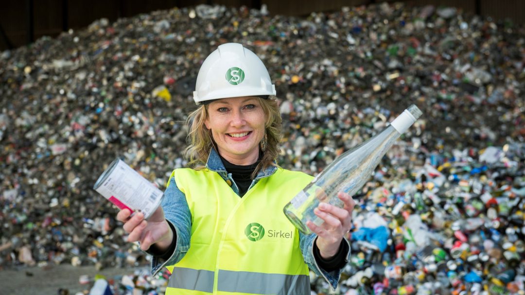 Gunhild-Solberg med refleksvest og vernehjelm som holder en metallboks og glassflaske. EN haug med sortert glass og metall ligger bak henne.