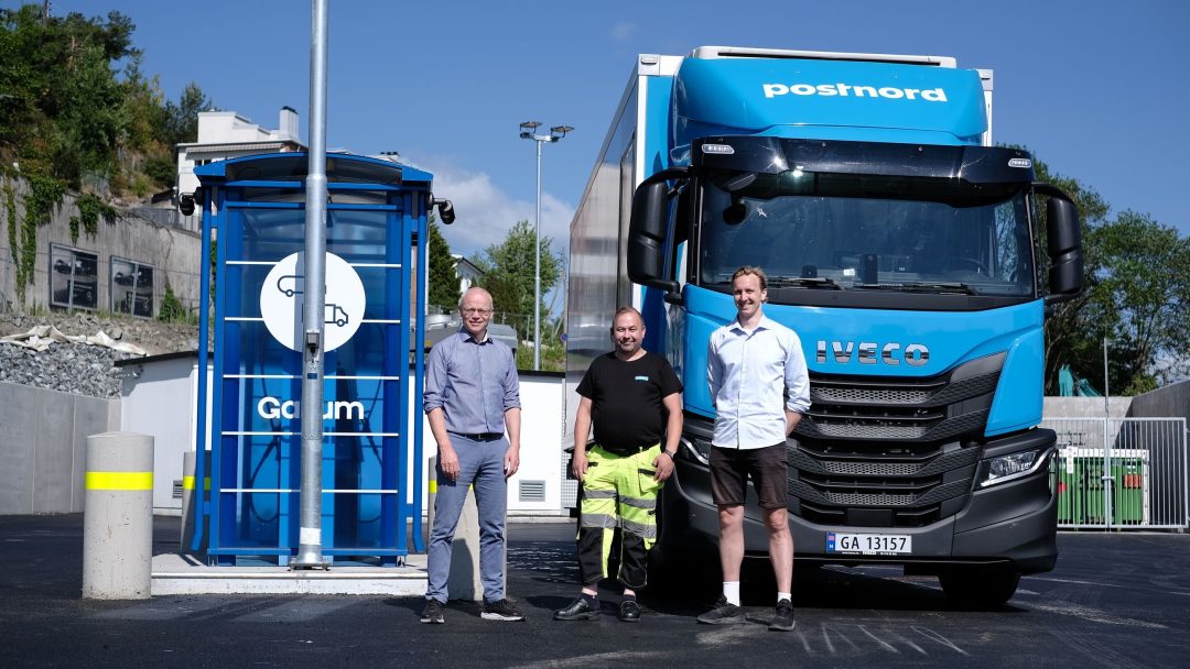 Tre menn står ved siden av en blå lastebil foran en energistasjon.
