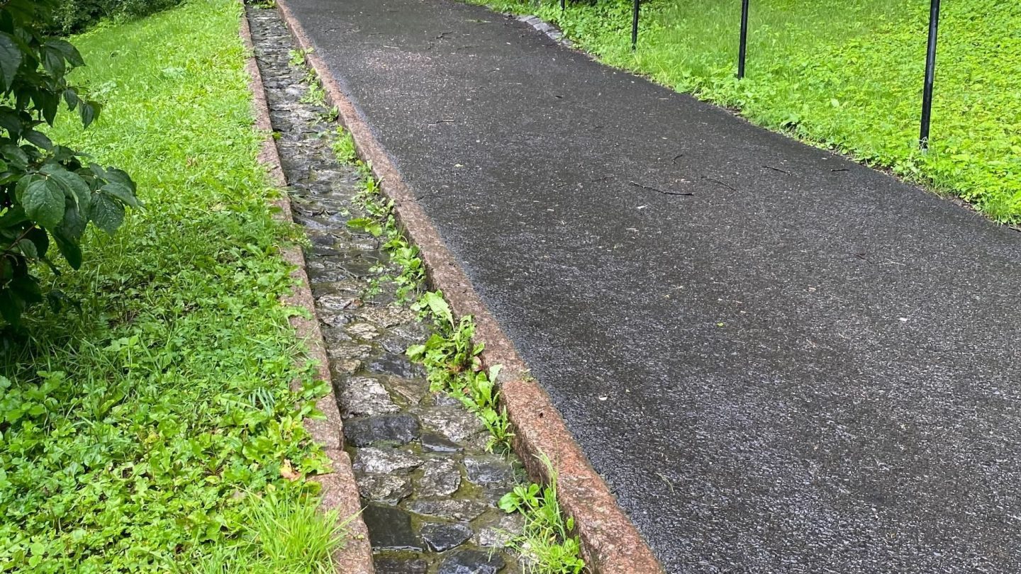 Et vanlig asfaltert gangvei i en nedoverbakke, ved siden av ser man en liten renne av stein som skal lede vannet ned i Iladalen park.