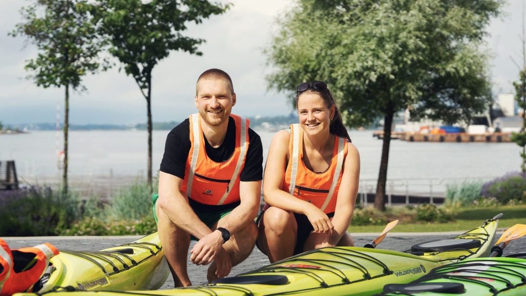 to personer på huk foran kajakker med vannet i bakgrunnen