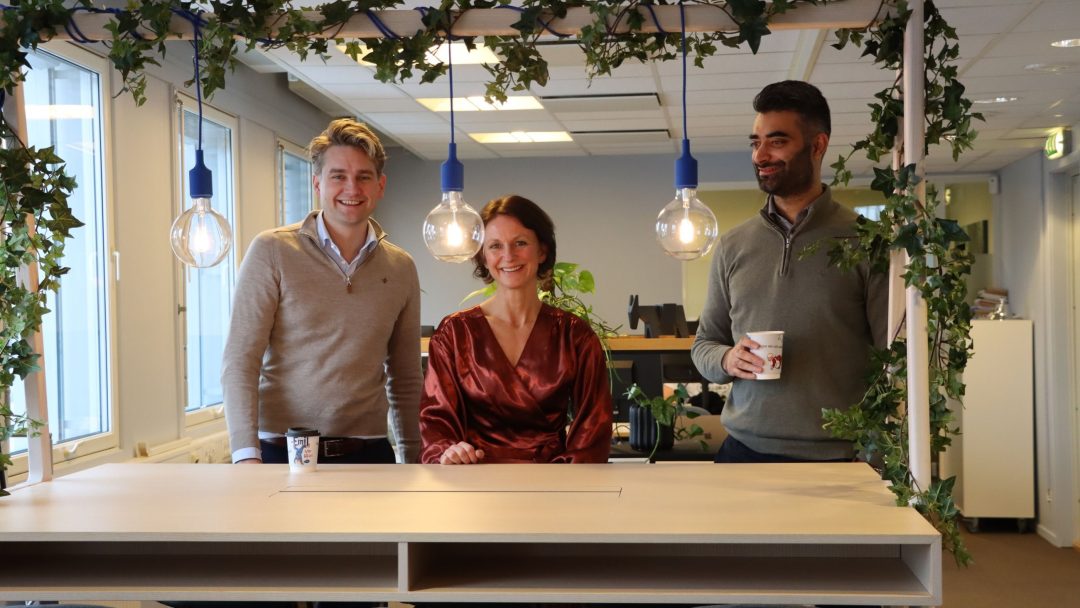Tre personer som står foran et bord på et kontor med kontormøbler.
