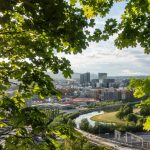 Bybilde innrammet av grønne blader, med en elv som slynger seg gjennom bygninger under en delvis overskyet himmel.