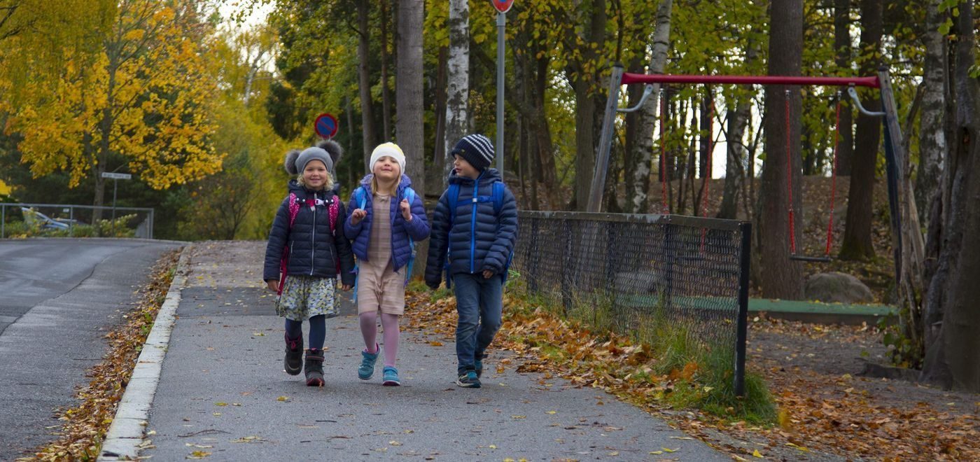 Tre barn på vei til skolen på høsten. Asfalt og gulgrønne trær rundt.