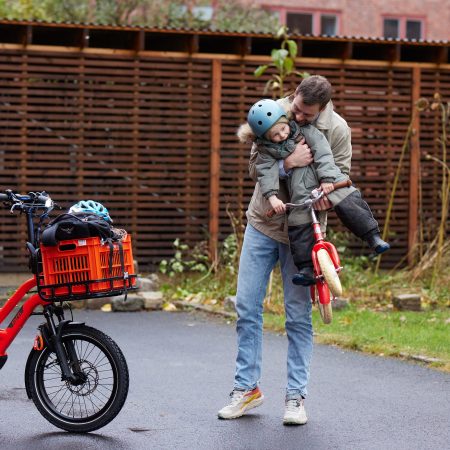 En far omfavner barnet sitt ved siden av en lastesykkel i nærheten av sykkelskur.