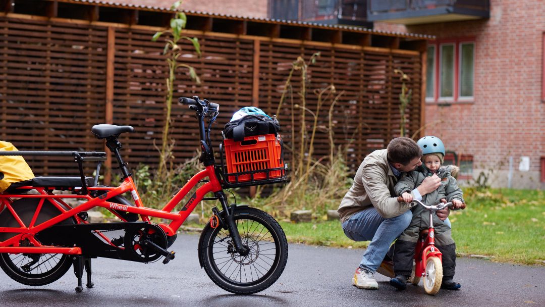 Voksen assisterer et barn på en liten sykkel ved siden av en parkert rød lastesykkel i en sykkelskur.