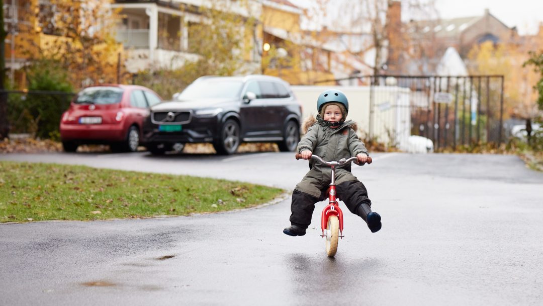 Et lite barn sykler på trehjulssykkel i en våt boliggate, på vei mot sykkelskur, iført hjelm og varme klær.
