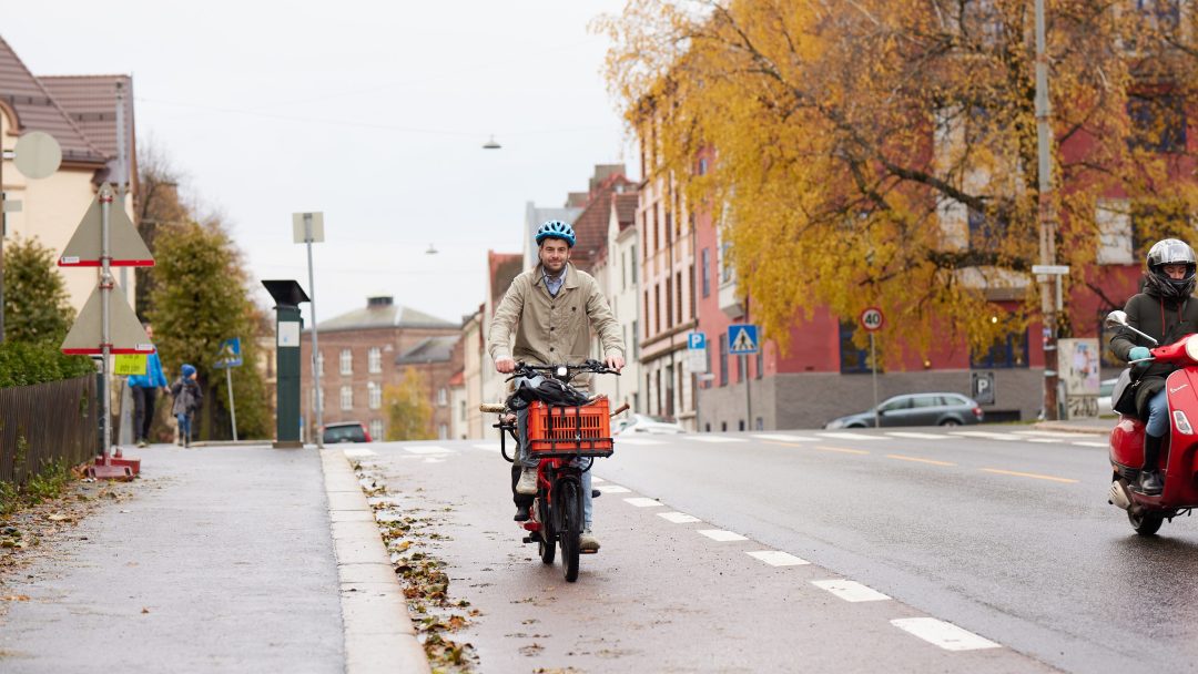 En person som kjører en lastesykkel på et bysykkelfelt ved siden av en gate med høsttrær i bakgrunnen, i nærheten av en sykkelskur.