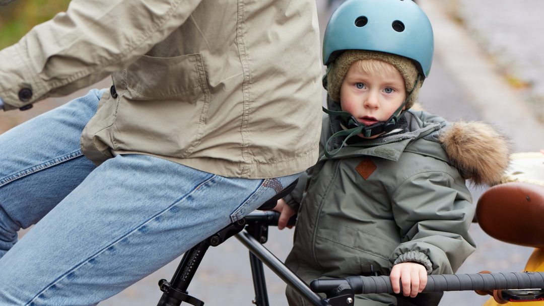 Et barn med blå hjelm sykler på sykkelfeste i nærheten av sykkelskur og ser til siden.