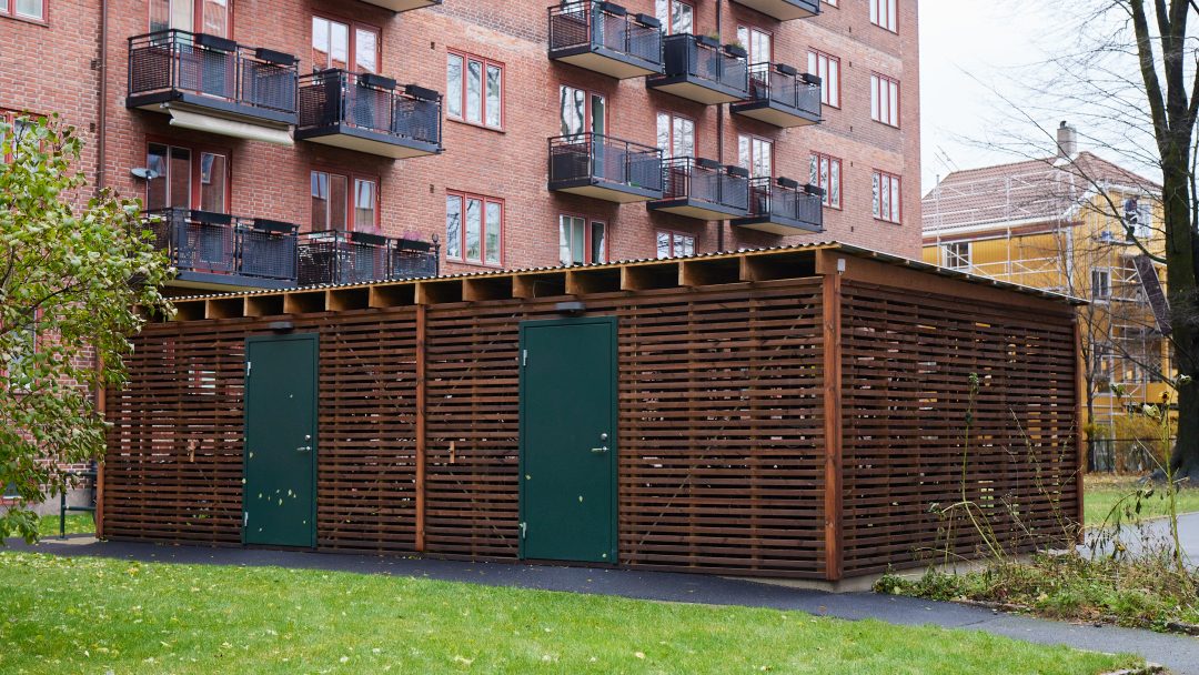 Tresykkelskur med grønne dører plassert i et boligområde med bygårder i bakgrunnen.