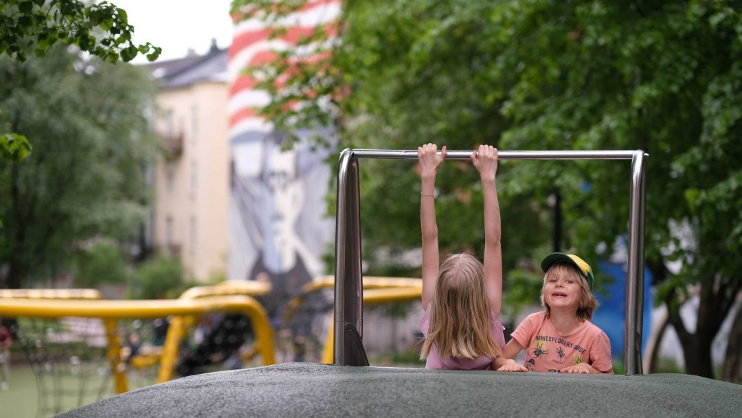 Iben henger i stativet, Aslak ser inn i kameraet. Vi ser gatekunst med Munch-motiv på bygning i bakgrunnen. lekeplasser