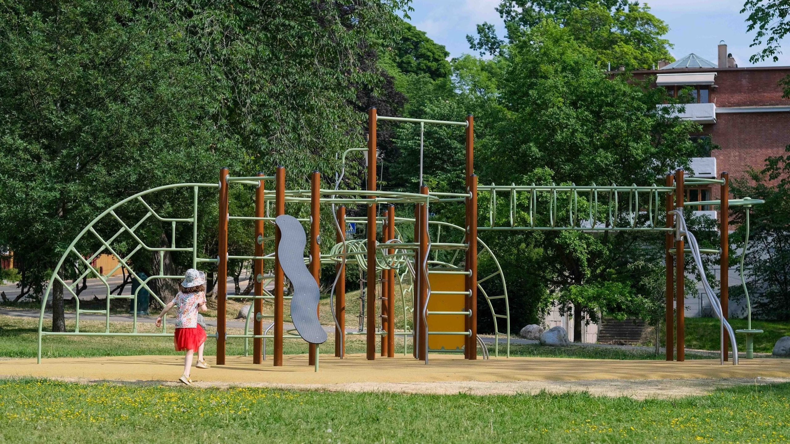 En liten jente på vei mot lekestativet i SKarpsnoparken. Grønt gress og frodige trær omringer lekestativet.