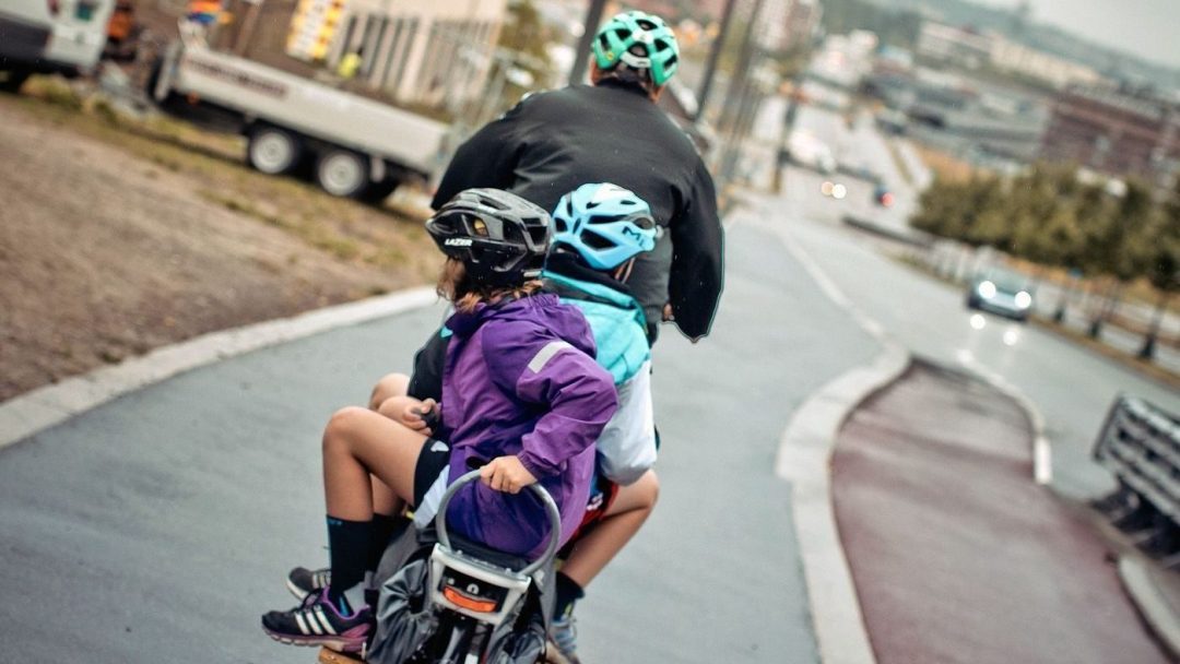 En som sykler med to barn bakpå. Bildet er tatt bakfra