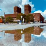 Mursteinsbygning med to tårn, med en klokke, reflekteres i en sølepytt. Trær og mennesker er synlige under en delvis overskyet himmel.