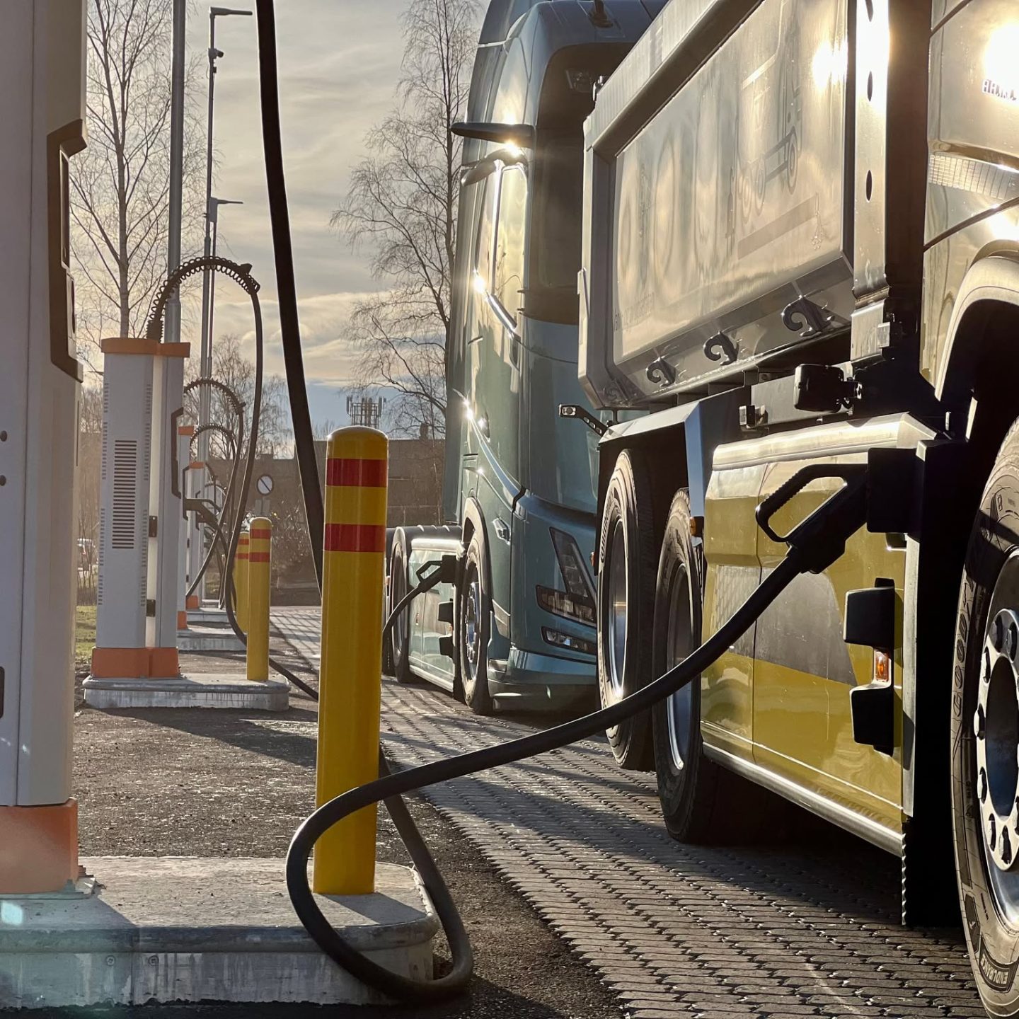 Under en klar blå himmel står tungtransport elektriske lastebiler i kø ved en ladestasjon, hver perfekt koblet sammen med solide kabler, klare til å starte veien videre.