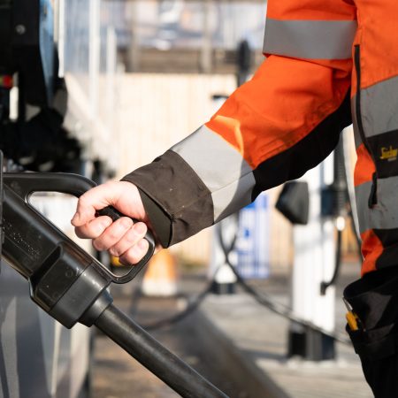 Person i en oransje sikkerhetsjakke som holder en ladeplugg for elektrisk kjøretøy, forbereder seg på å lade et kjøretøy.