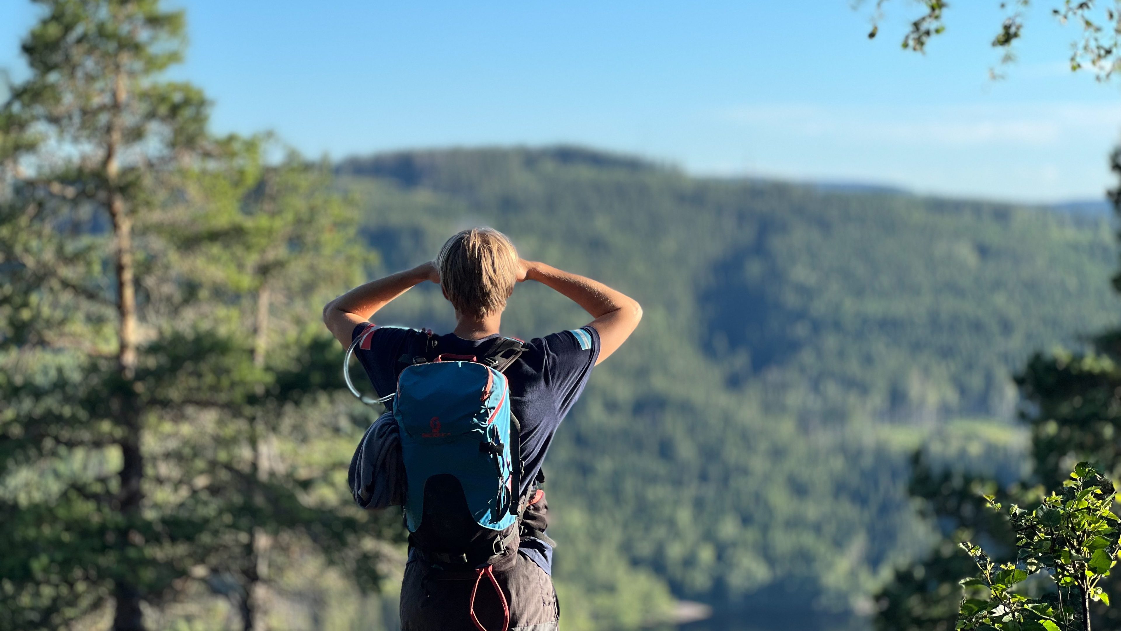Person med ryggsekk som står og ser på et skogkledd landskap og innsjø under en klar blå himmel, og nyter den givende utsikten etter en spennende topptur.