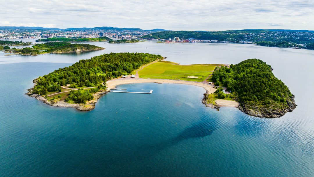 En luftfoto avslører en frodig grønn øy med teltskoger og åpne marker, omgitt av glitrende vann. En liten brygge strekker seg innbydende inn i de asurblå bølgene. I det fjerne ligger et byområde under en delvis overskyet himmel.