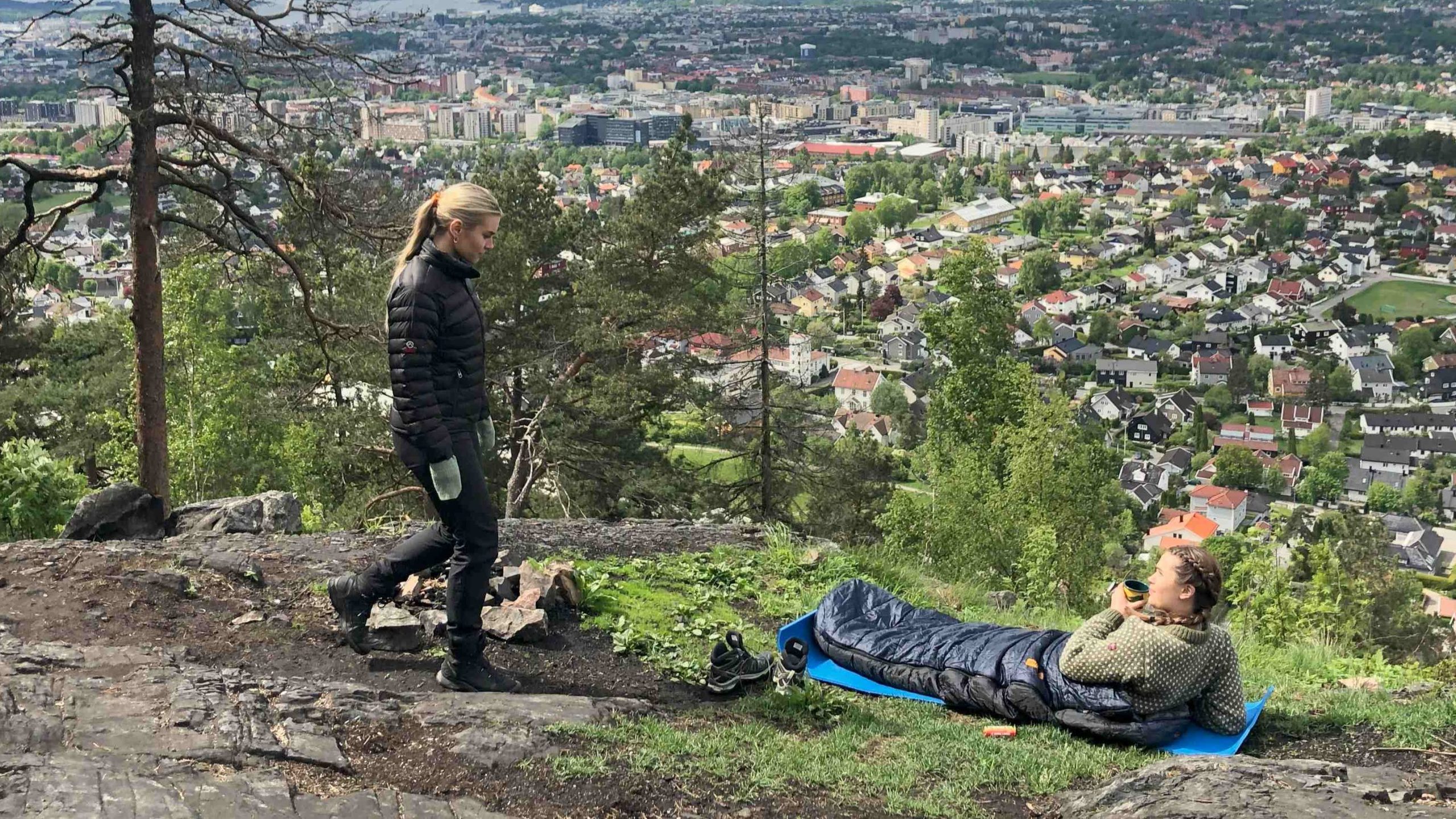 To personer på en steinete høyde med utsikt over et bybilde, den ene står i friluftsutstyr ved siden av telten, mens den andre hviler på en blå campingmatte i en sovepose. En overskyet himmel ruver over.