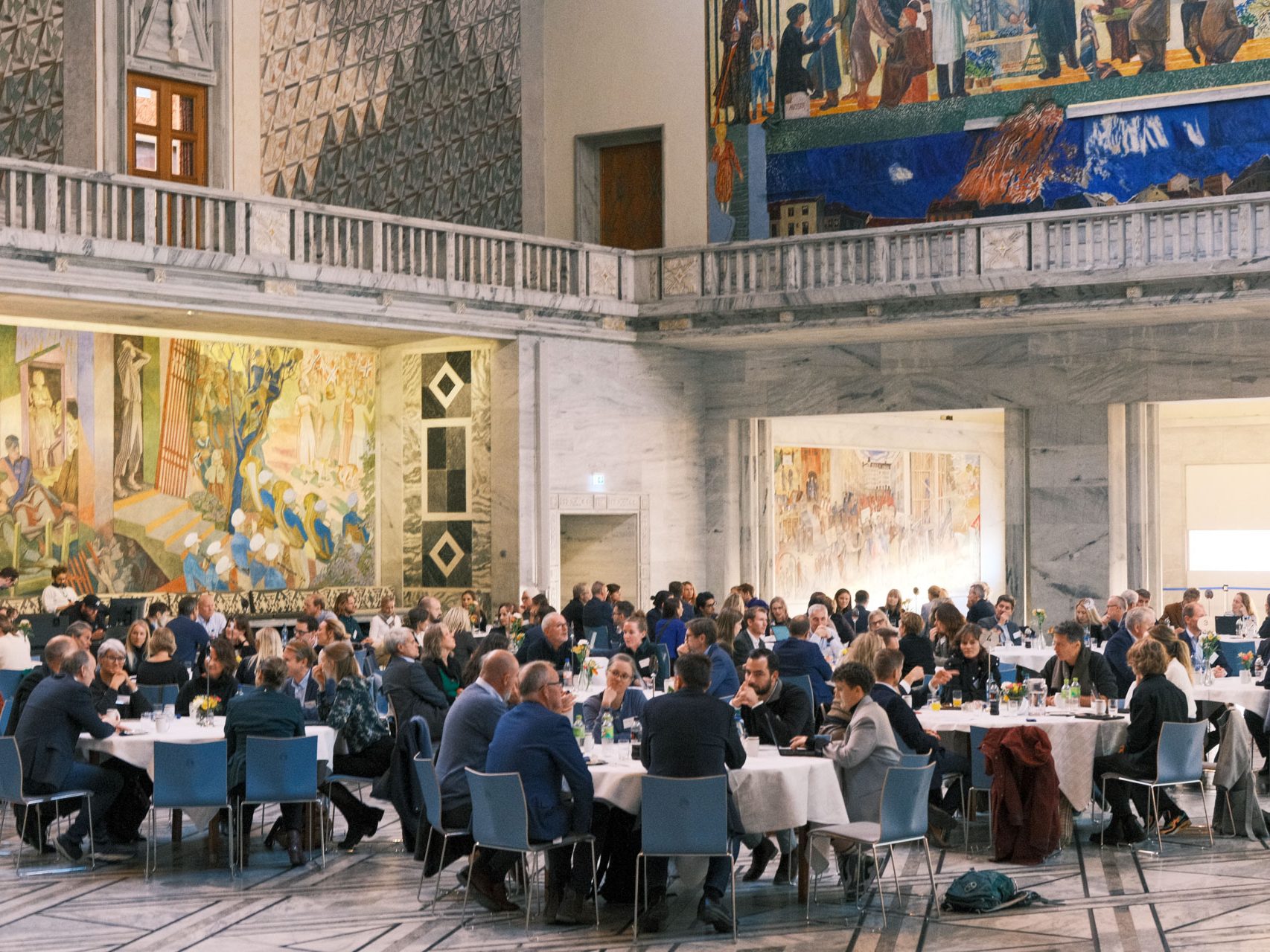 Folk satt ved runde bord i en stor, utsmykket hall med fargerike veggmalerier på veggene.