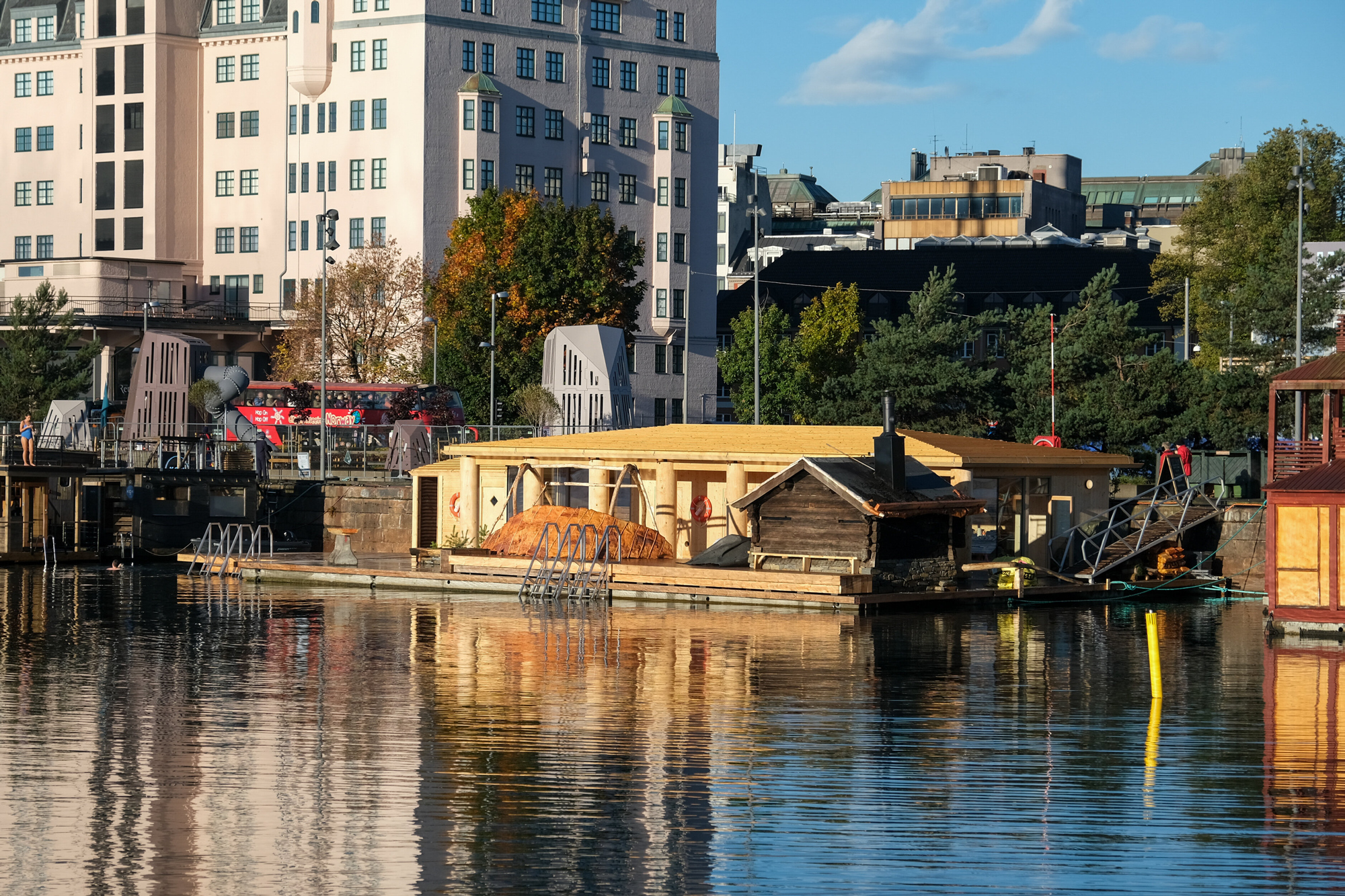En flytende badstu-konstruksjon med en liten trehytte ved havnen, som speiler seg i vannet foran bybygningene.