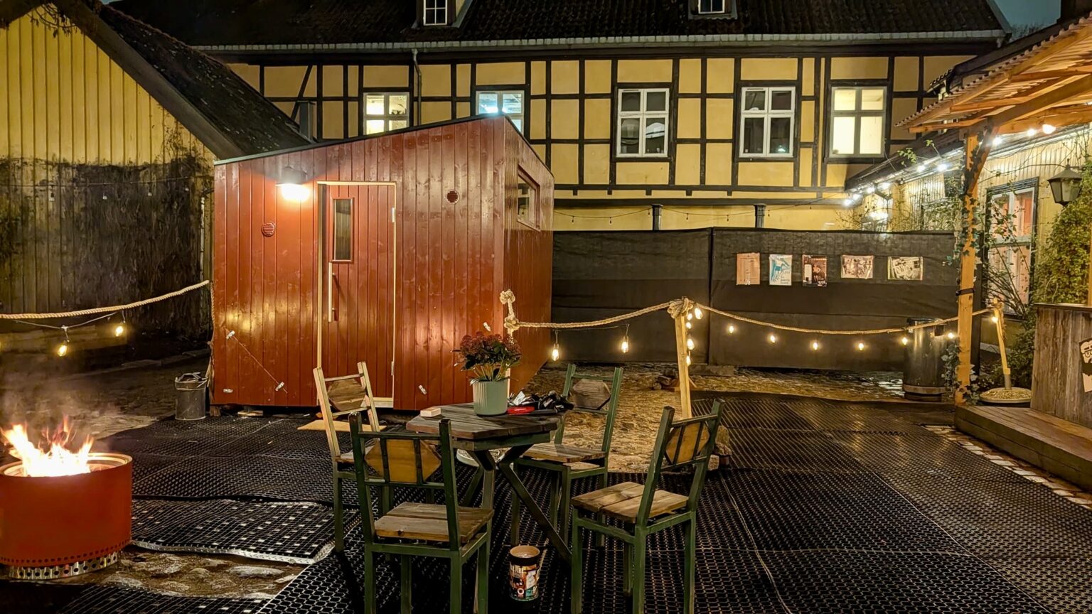 Uteplass med trestoler og bord, en potteplante som borddekorasjon, lyskjeder, et rødt skur og et bålsted om kvelden. I bakgrunnen står en gul og brun bindingsverksbygning ved siden av en koselig badstu, noe som skaper en innbydende atmosfære.