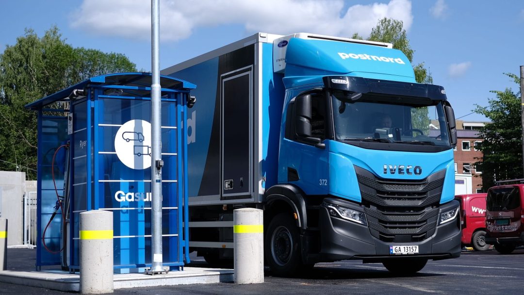 En blå lastebil fra PostNord står parkert ved en fyllestasjon for biogass