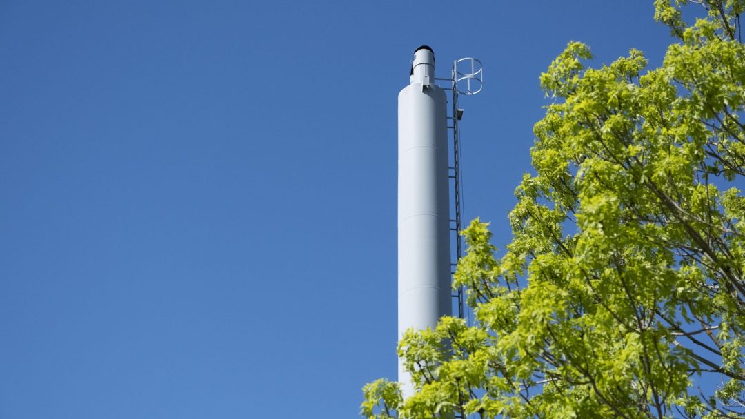 En høy, hvit industriskorstein med stige står mot en klarblå himmel ved siden av grønne, løvrike tregrener.