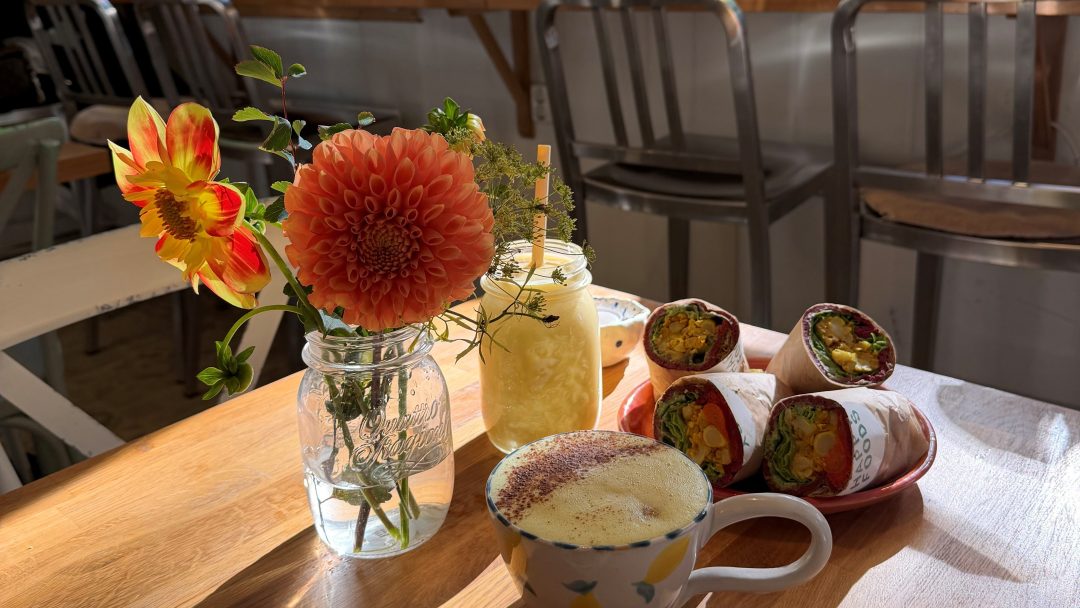 Bilde fra Happy Foods inne i lokalet med sol igjennom vinduene, blomster på bordet, en kaffekopp og noen wraps.