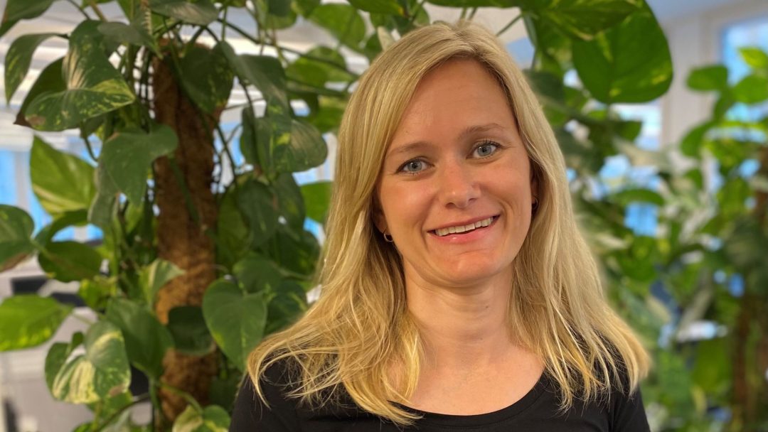 Person med langt blondt hår i svart skjorte som står grasiøst foran store innendørs planter, og omfavner roen til nullutslippssoner.