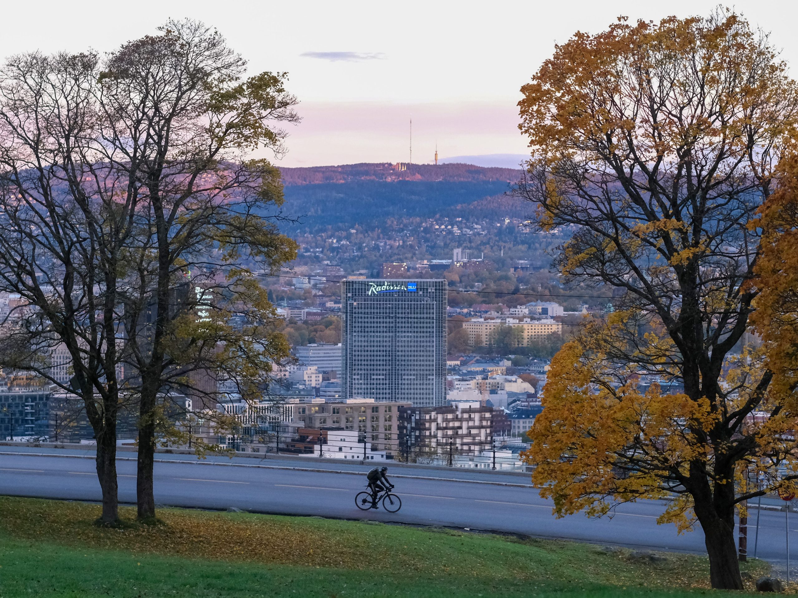 En person sykler på en asfaltert vei kantet av høsttrær, med et bylandskap og åser i bakgrunnen.