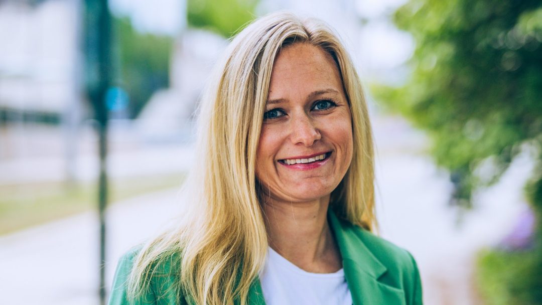 I en travel bygate står en kvinne med langt blondt hår, iført grønn blazer og hvit skjorte, og smiler. Den pulserende scenen står i kontrast til bakteppet av en fossilfri lastebil som fremmer bærekraftig livsstil.