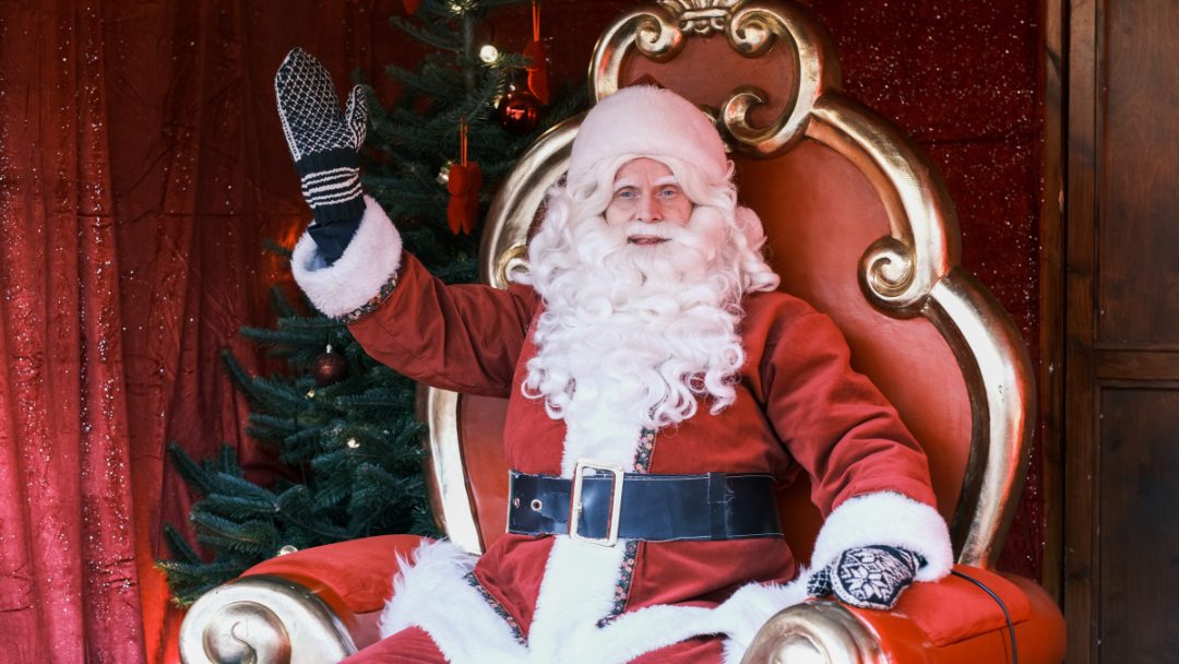 Julenissen sitter på en rød og gullfarget trone og vinker, mens et pyntet juletre - og en stabel brukte julegaver - glitrer i bakgrunnen.