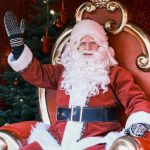 Julenissen sitter på en rød og gullfarget trone og vinker, mens et pyntet juletre - og en stabel brukte julegaver - glitrer i bakgrunnen.