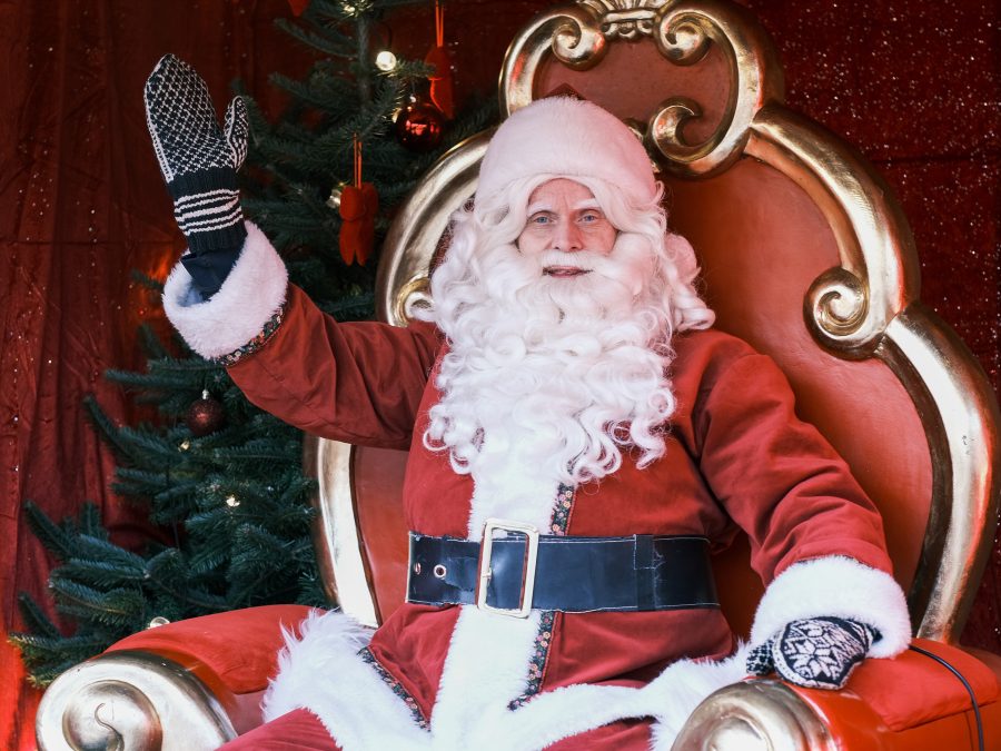 Julenissen sitter på en rød og gullfarget trone og vinker, mens et pyntet juletre - og en stabel brukte julegaver - glitrer i bakgrunnen.