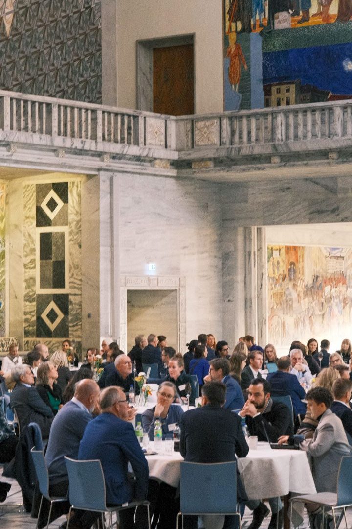 Folk satt ved runde bord i en stor, utsmykket hall med fargerike veggmalerier på veggene.