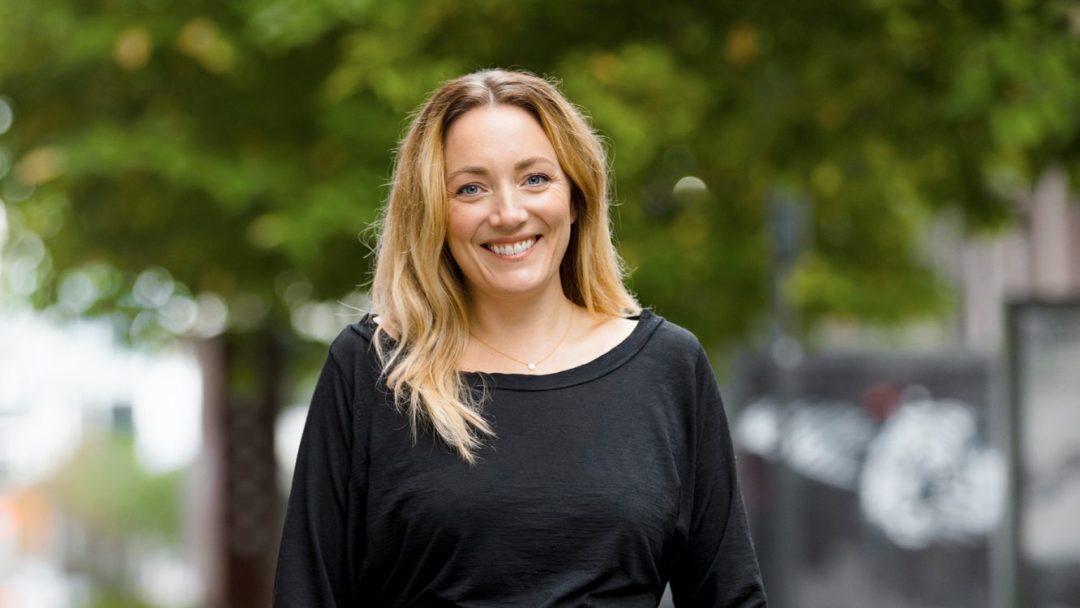 En person står utendørs i en allé og smiler blant de solplettede bladene, iført en svart topp og olivengrønne bukser med hendene i lommene, uberørt av den lette overvannssamlingen på fortauet.