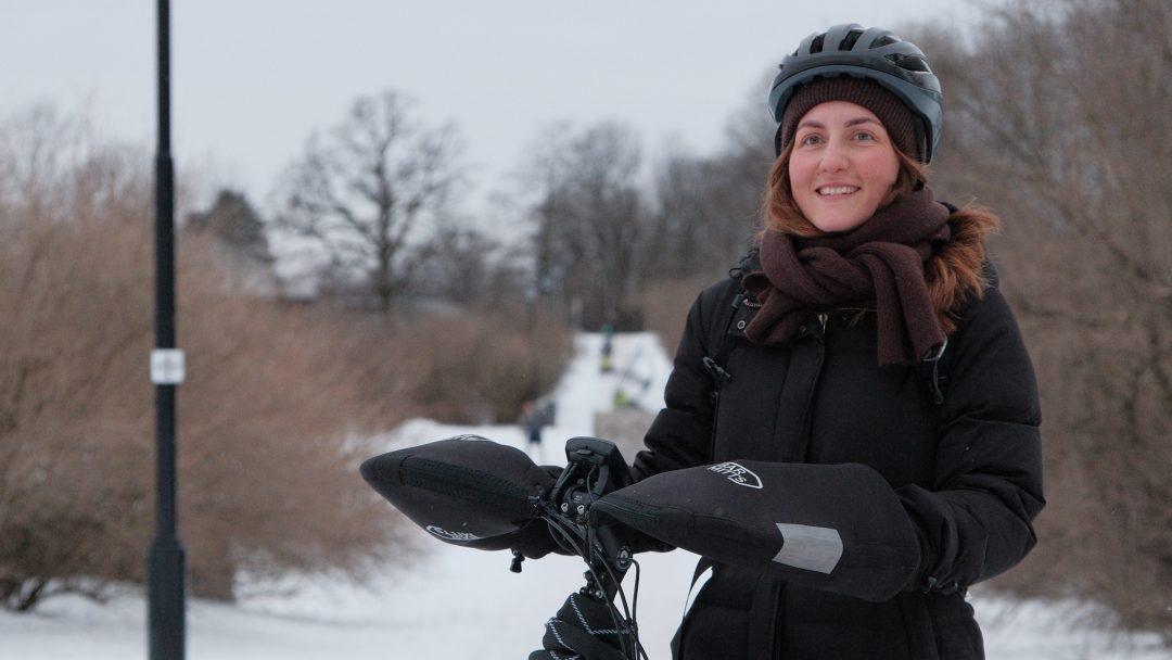 En person med hjelm og vinterfrakk står med en sykkel på en snødekt sti, omgitt av løvløse trær på en overskyet dag.