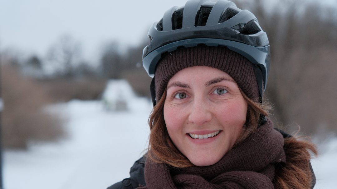En kvinne med sykkelhjelm, brunt skjerf og brun lue smiler utendørs en snørik vinterdag.