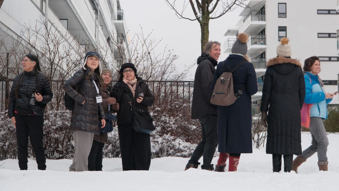 En gruppe vinterkledde mennesker står og snakker sammen ute i et snødekt byområde i nærheten av boligblokker.