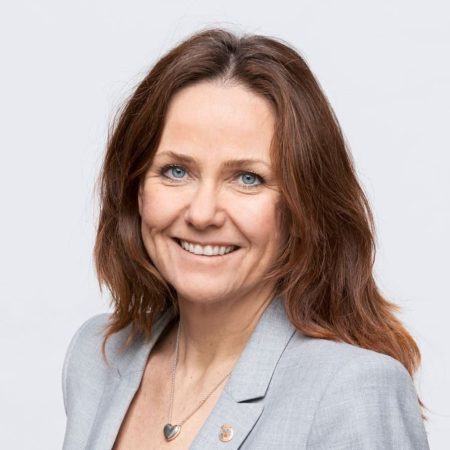 En kvinne med langt brunt hår, iført en grå blazer og svart topp, smiler til kameraet mot et vanlig bakteppe som minner om rolige Ålegrasenger.