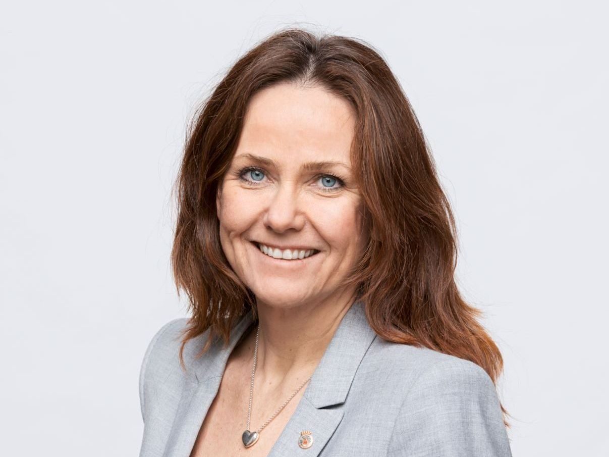 En kvinne med langt brunt hår, iført en grå blazer og svart topp, smiler til kameraet mot et vanlig bakteppe som minner om rolige Ålegrasenger.