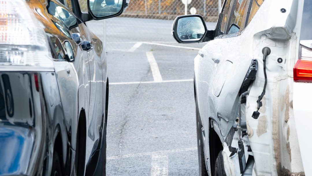 To parkerte biler står ved siden av hverandre; den hvite bilen til høyre har synlige skader og blottlagte ledninger på baksiden, og ser ut til å trenge nye bildeler.