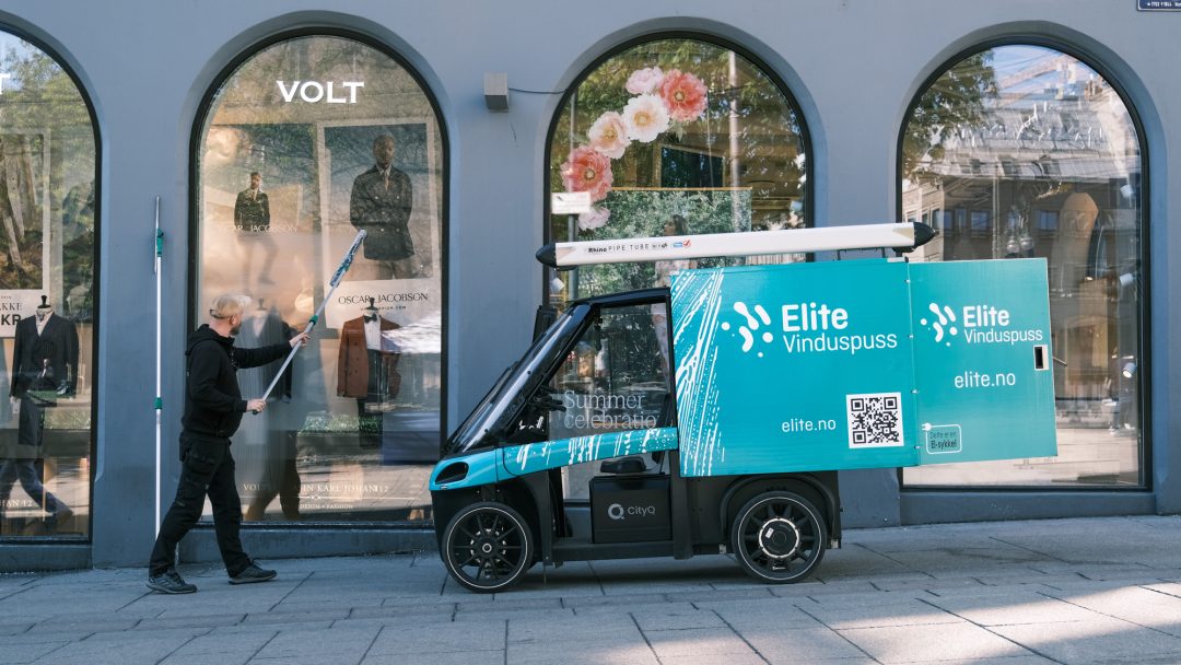 En vinduspusser bruker et verktøy med langt skaft på et butikkvindu ved siden av en liten lastesykkel av merket Elite Vinduspuss, mens rengjøringsbilen deres står parkert i en travle bygate.