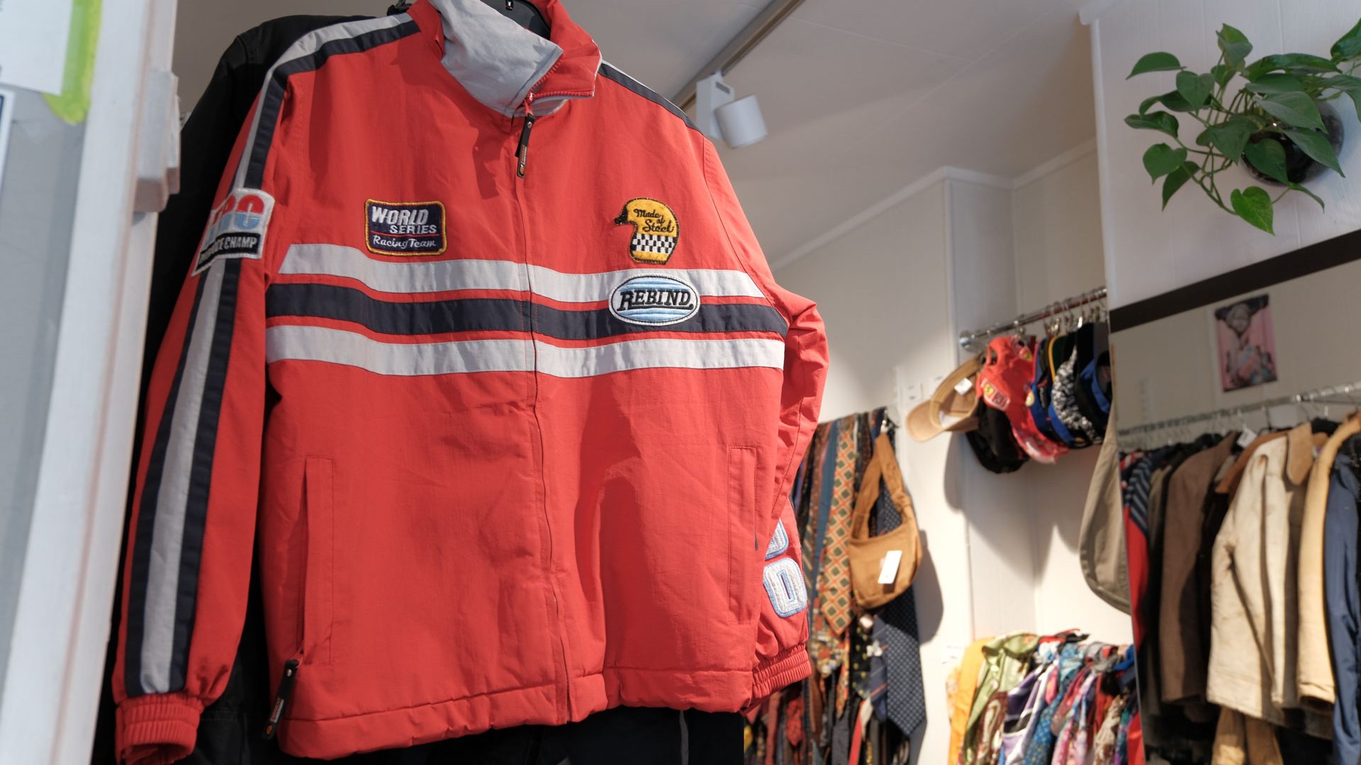 Klær og kvalitet. En rød jakke i racingstil med lapper og striper, kjent for sin kvalitet, henger utstilt i en klesbutikk, omgitt av en rekke andre plagg og tilbehør.