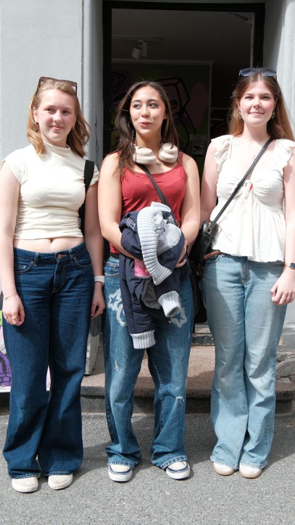 Tre unge kvinner står side om side på fortauet, kledd i uformelle antrekk med jeans og topper som fremhever klær og kvalitet, og poserer for kameraet foran inngangen til en bygning.