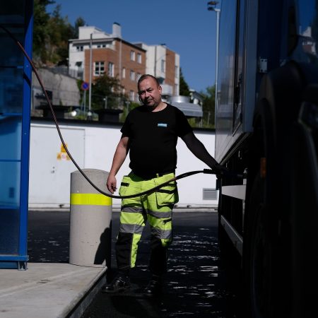 En person i synlige bukser fyller drivstoff på en biogassstasjon, mens han holder slangen med én hånd. Bygninger og trær danner bakteppet.