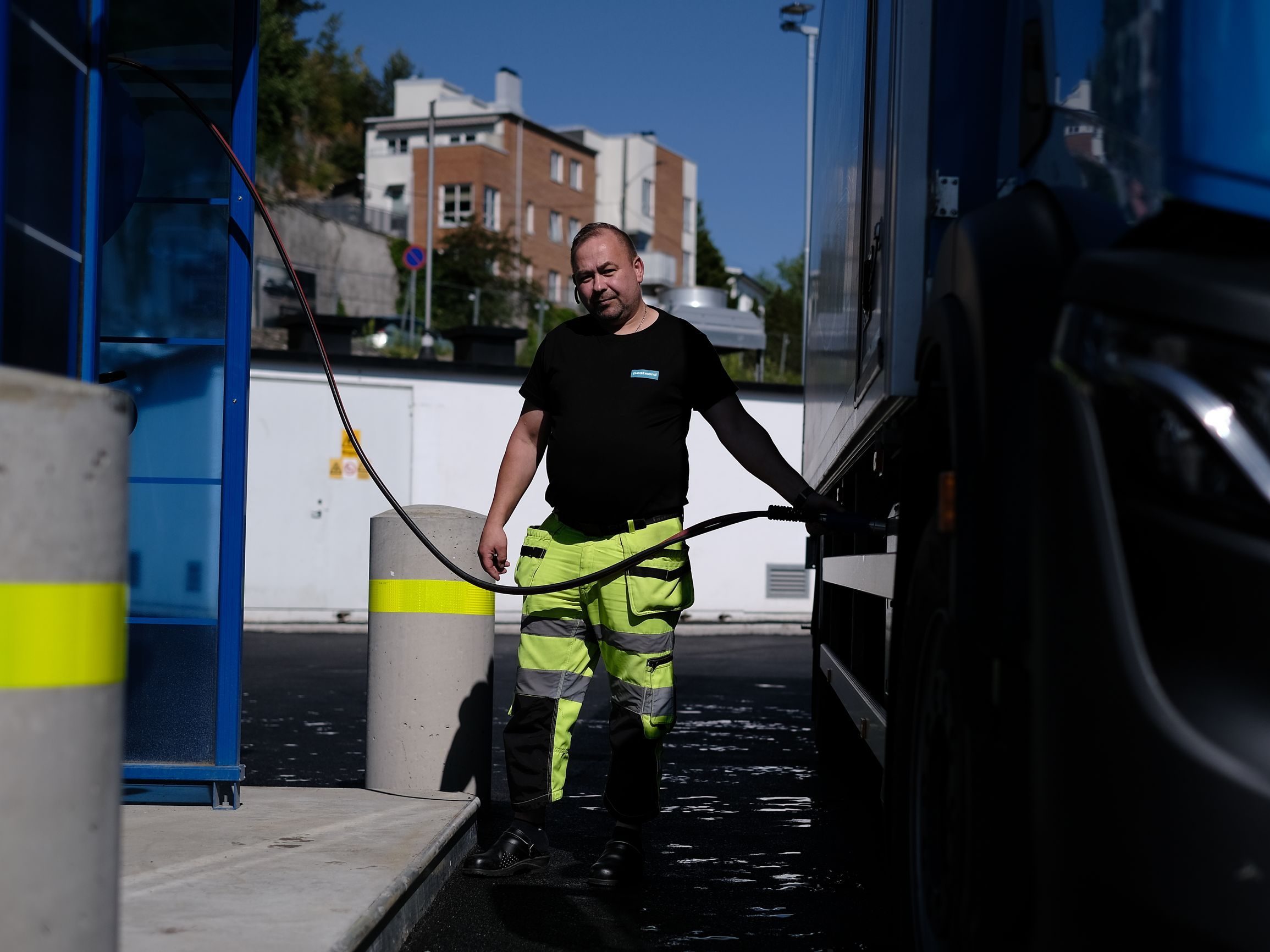En person i synlige bukser fyller drivstoff på en biogassstasjon, mens han holder slangen med én hånd. Bygninger og trær danner bakteppet.